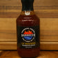 A bottle of Crooked Creek Barbecue Lip Smacking Barbecue Sauce, featuring a black cap and black label with red and yellow text, set on a wooden surface against a wooden background.