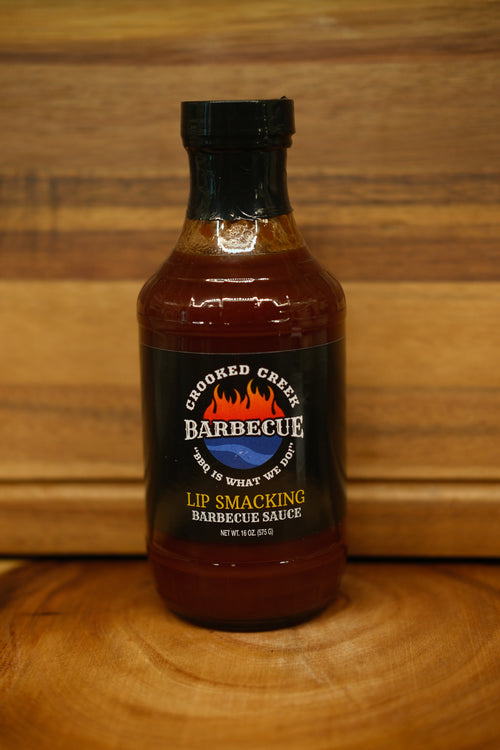 A bottle of Crooked Creek Barbecue Lip Smacking Barbecue Sauce, featuring a black cap and black label with red and yellow text, set on a wooden surface against a wooden background.