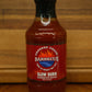 A bottle of Chopped Creek Barbecue Slow Burn Barbecue Sauce, featuring a red label and black cap, set on a wooden surface against a wooden background.