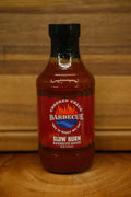 A bottle of Chopped Creek Barbecue Slow Burn Barbecue Sauce, featuring a red label and black cap, set on a wooden surface against a wooden background.