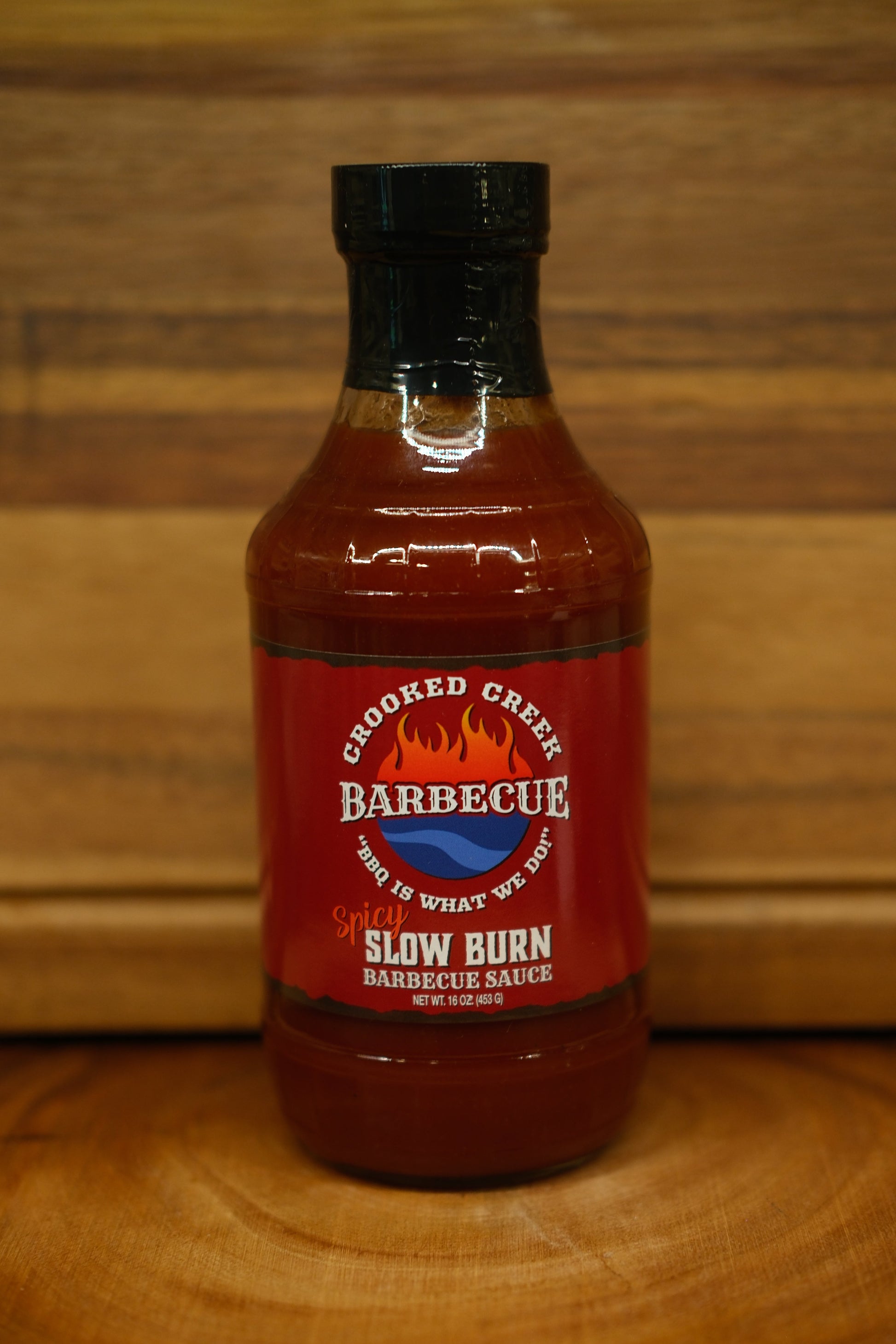 A bottle of Chopped Creek Barbecue Slow Burn Barbecue Sauce, featuring a red label and black cap, set on a wooden surface against a wooden background.