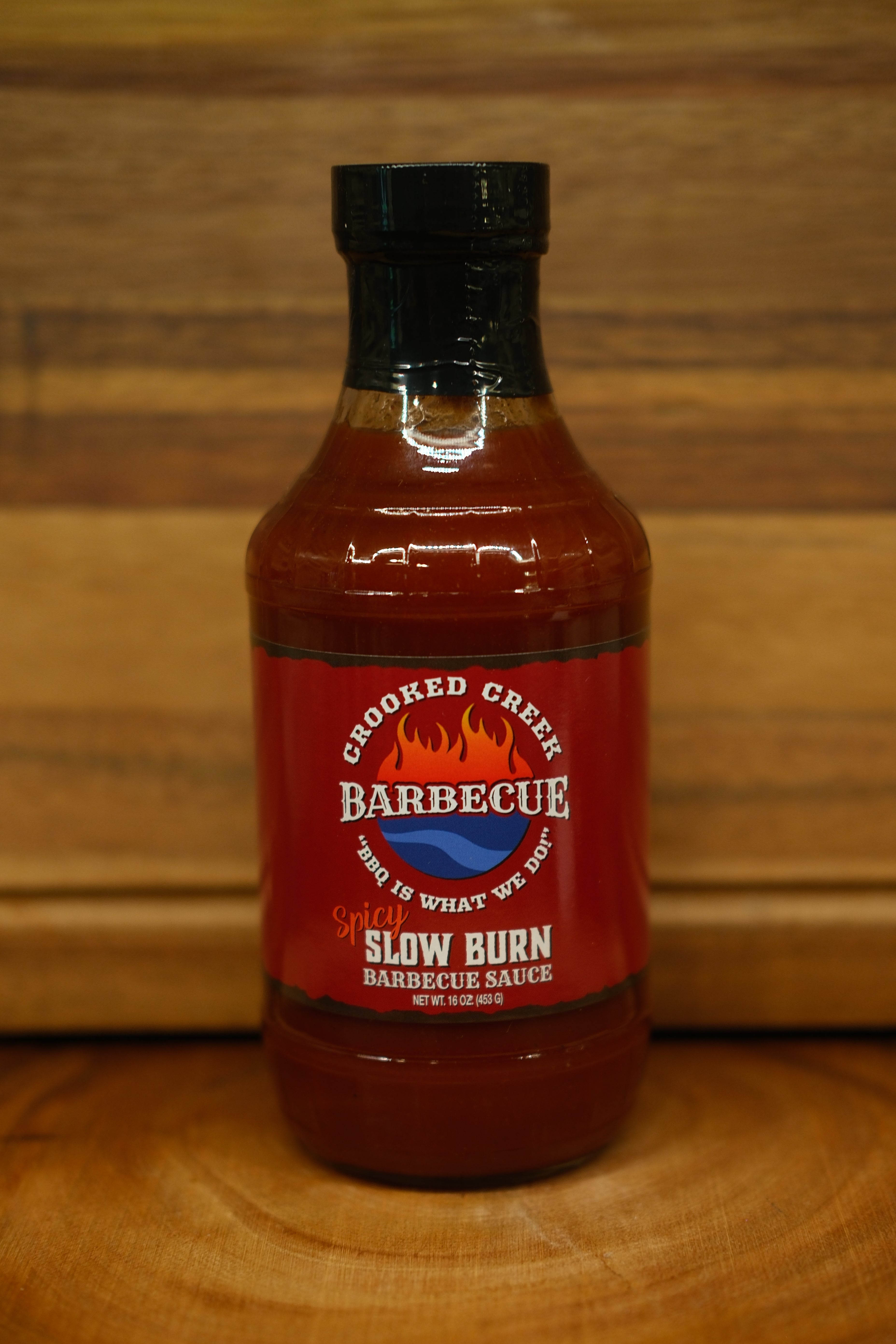 A bottle of Chopped Creek Barbecue Slow Burn Barbecue Sauce, featuring a red label and black cap, set on a wooden surface against a wooden background.